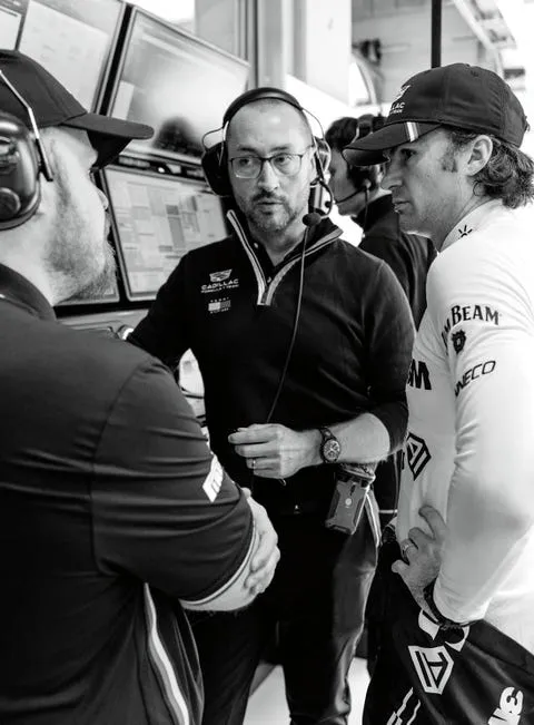 Three professionals discussing strategy in a motorsport pit area, with screens displaying data in the background.