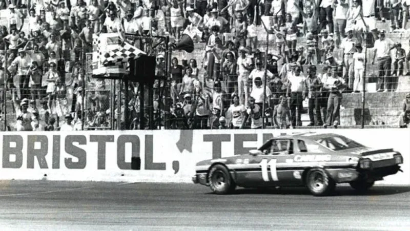 Race car number 11 driving past the Bristol Motor Speedway with cheering fans in the stands.