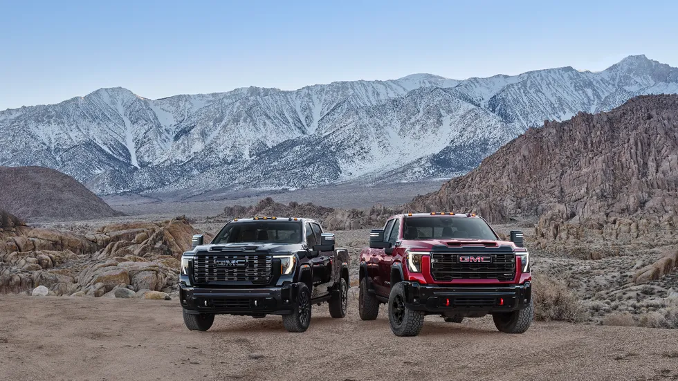 Two GMC trucks parked on rocky terrain with mountains in the background under a clear blue sky.