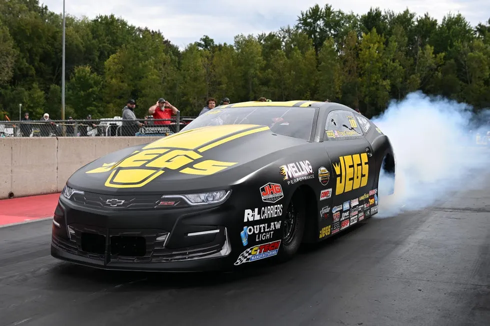 Black and yellow drag racing car emitting tire smoke on a racetrack with spectators in the background.